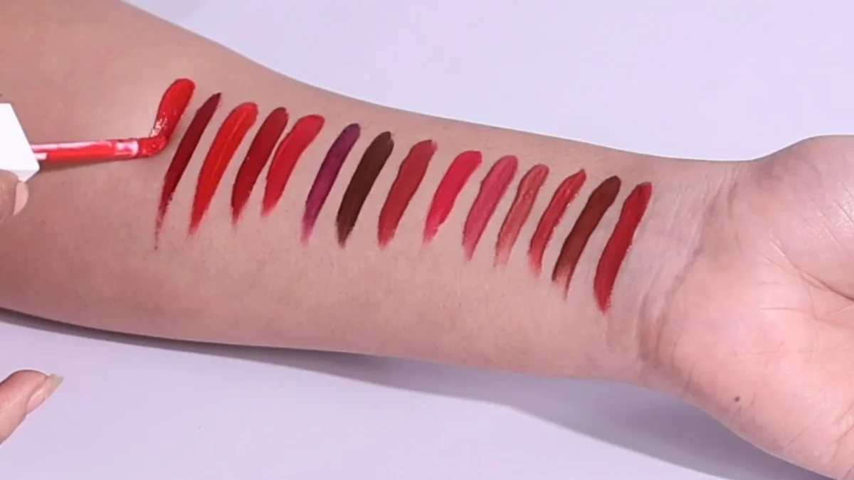 Close-up of a forearm with a row of red, rose and brown liquid lipstick swatches being applied with a doe-foot applicator on a lilac background, a beauty UGC shot that often needs longer listing usage than ad usage