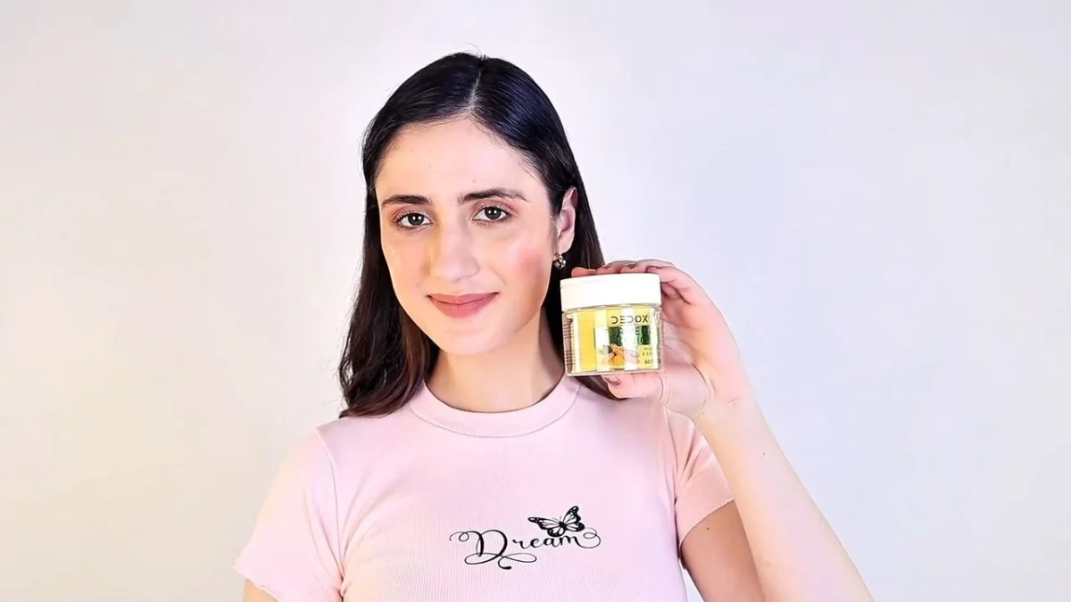 Indian woman creator in a pink t-shirt holding a small skincare jar beside her face against a plain light wall, a talking-to-camera setup that needs usage rights in the creator content brief