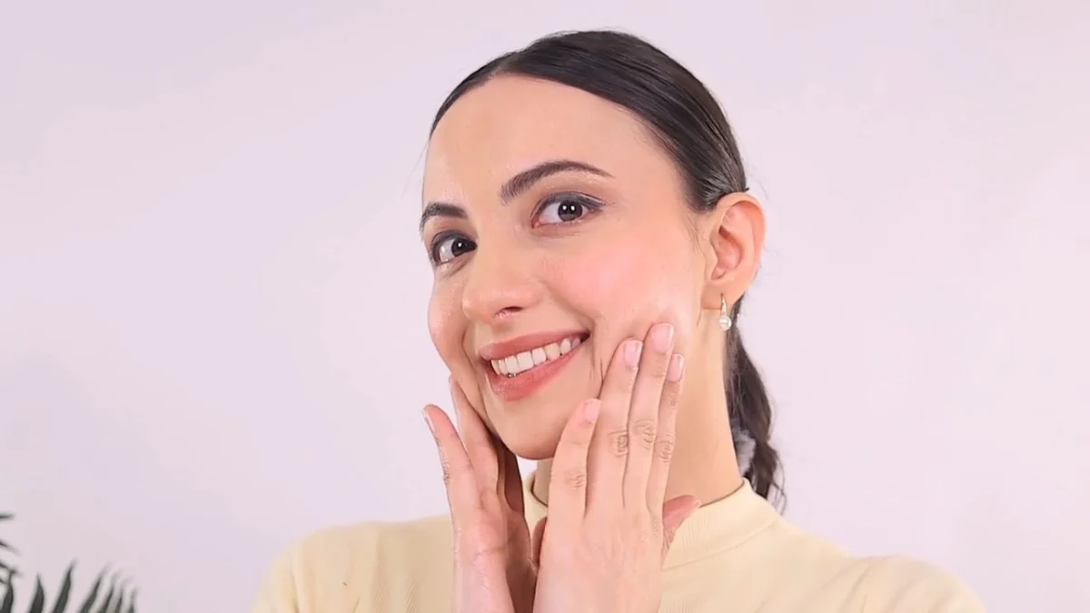 Indian woman creator with slicked-back hair smiling as she presses cream into her cheeks with both hands against a soft white backdrop, a close application beat listed in the UGC shoot brief