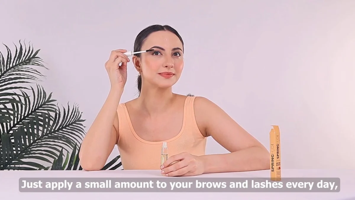 Indian woman creator in a peach top applying a brow serum with a spoolie at a white table beside a palm plant, with a subtitle line burned into the frame, a demo shot where creator content feedback should focus on product handling