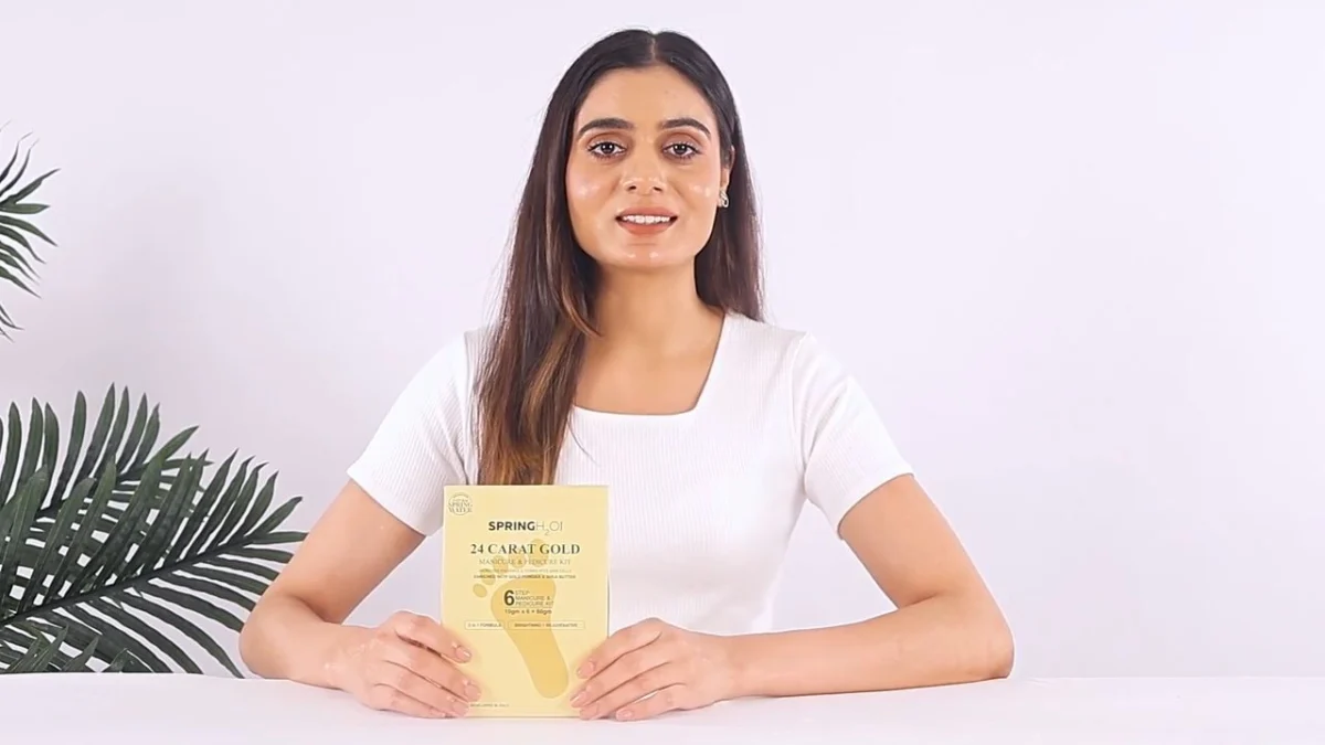 Indian woman creator in a white t-shirt seated at a white table holding a skincare product pack toward the camera on a bright home-style set, filmed from a UGC creator brief template