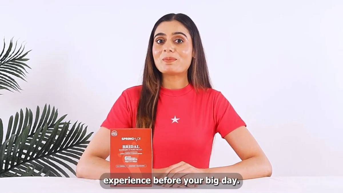 Indian woman creator in a red t-shirt seated at a white desk with plants, speaking directly to camera with a skincare product beside her, a talking-head testimonial UGC ad concept