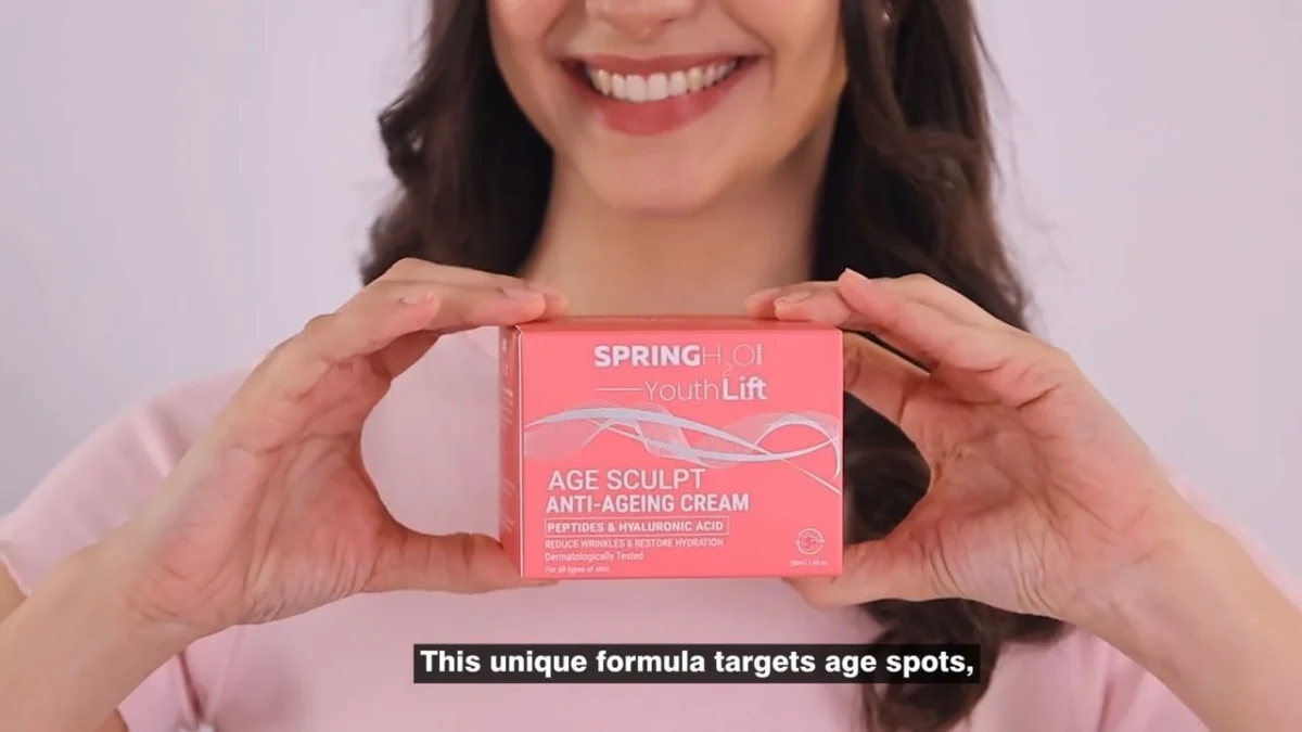 Smiling Indian woman creator holding an unopened skincare cream box up to the camera on a soft pink set with a burned-in caption line at the bottom, the kind of first cut that UGC video revisions feedback is written against