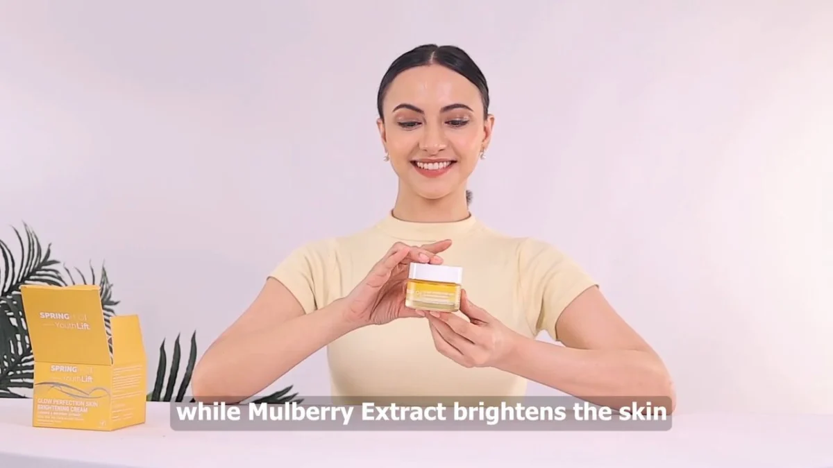 A woman handles an open skincare jar at a studio table while explaining a product on camera