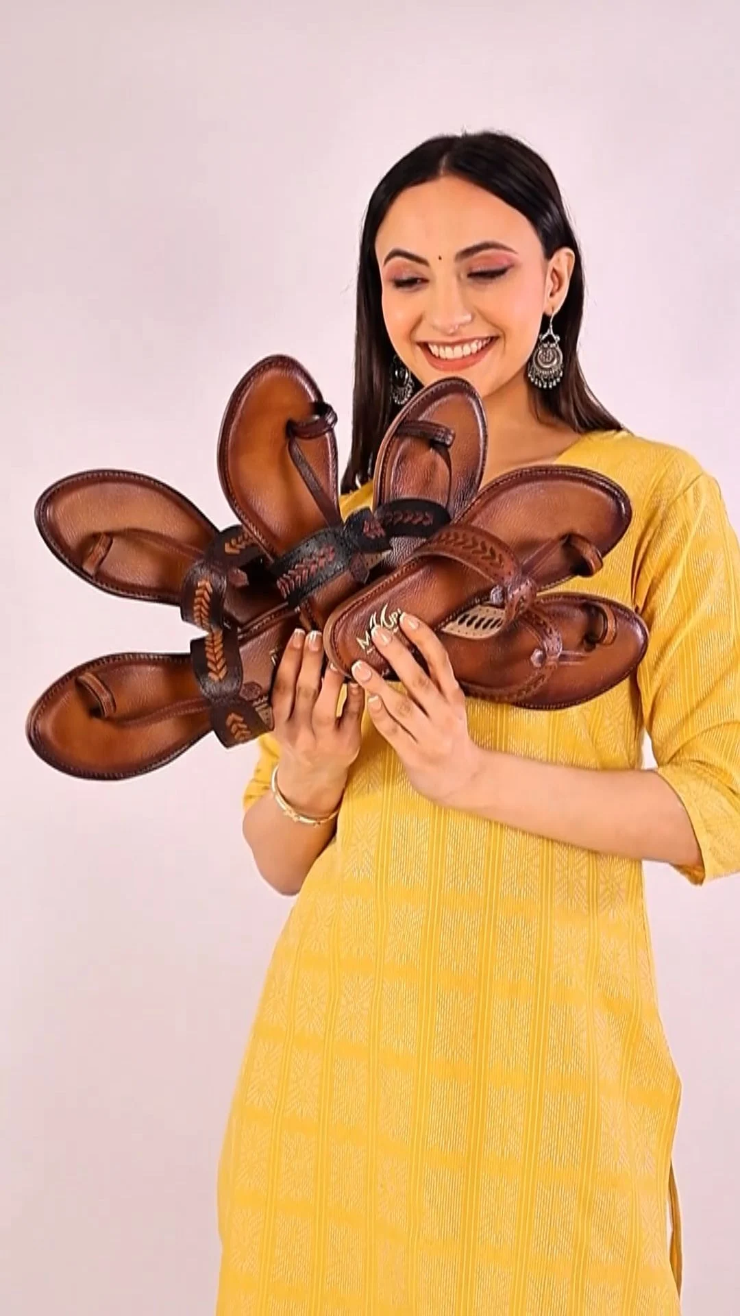 Smiling Indian woman in a yellow kurta and silver earrings holds a fan of brown leather sandals toward the camera in a bright studio, a creator product demo whose usage scope and duration sit in the UGC contract checklist