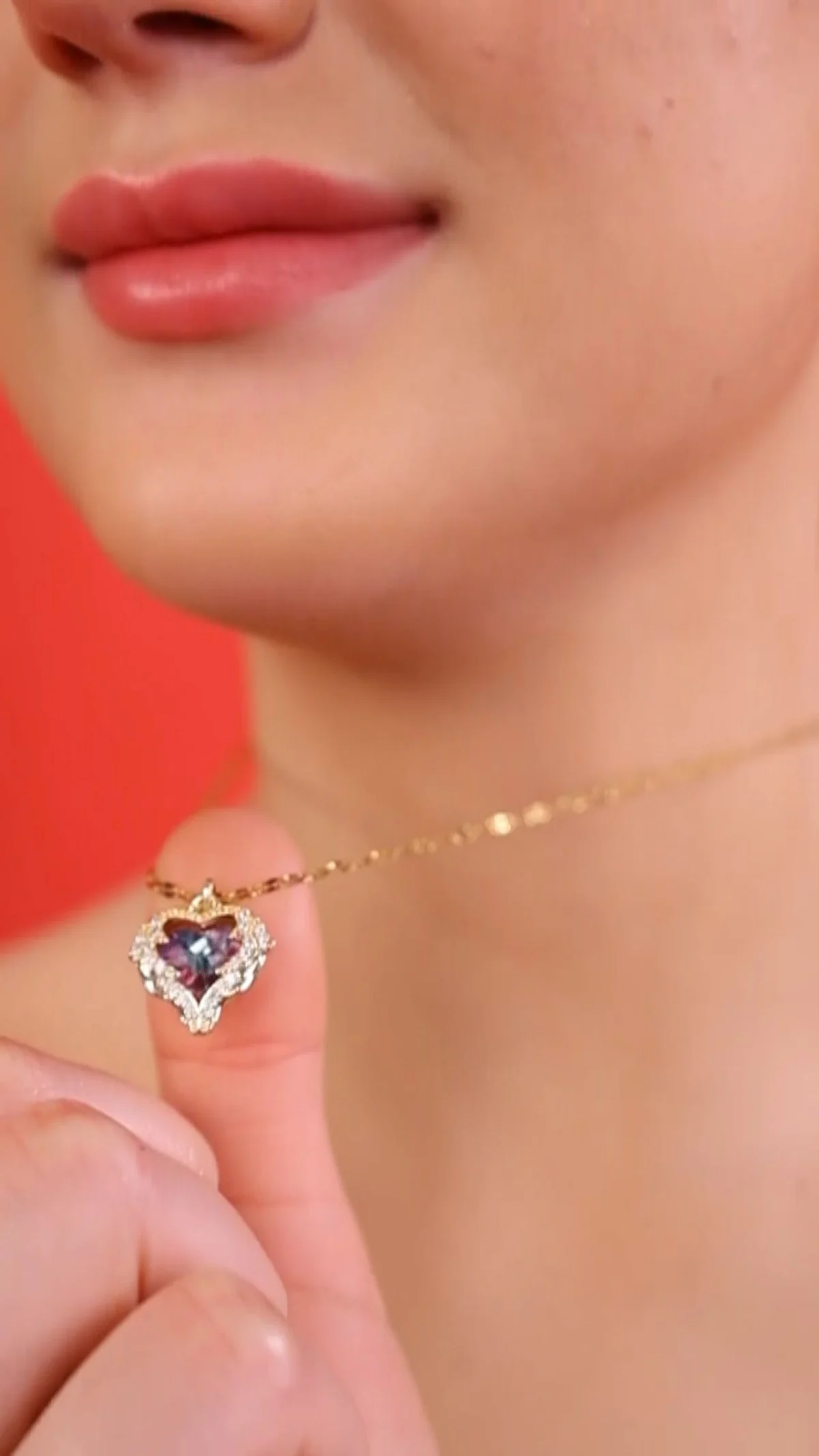 Close-up of an Indian model's chin and neck in front of a red backdrop as she holds a delicate gold chain with a heart-shaped pendant, jewellery detail shot that still needs model exclusivity terms and a release