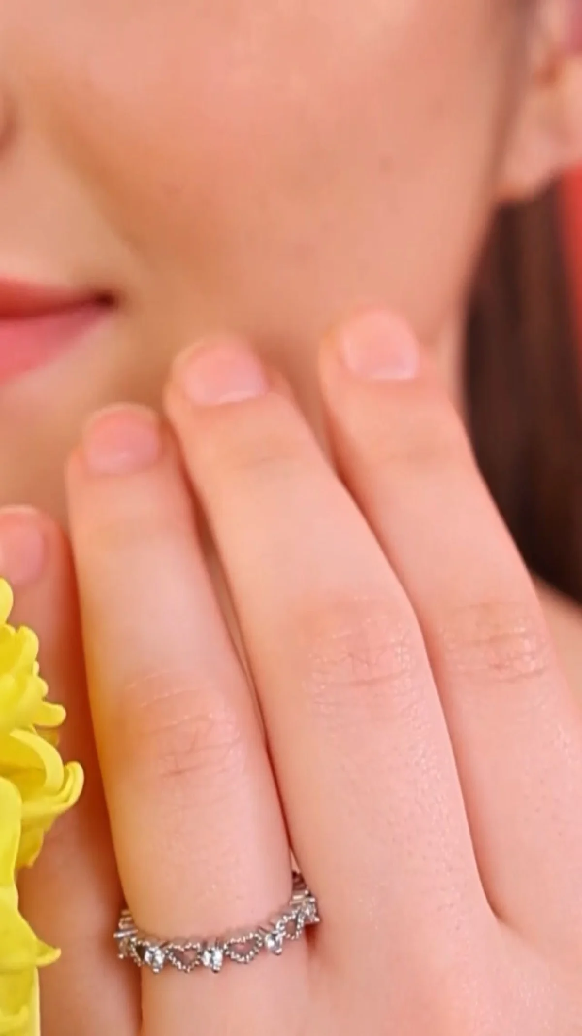 Close-up of a woman's hand with clean nude nails wearing a delicate silver ring near her lips, beside yellow flowers, showing the hand and nail detail jewellery casting teams look for