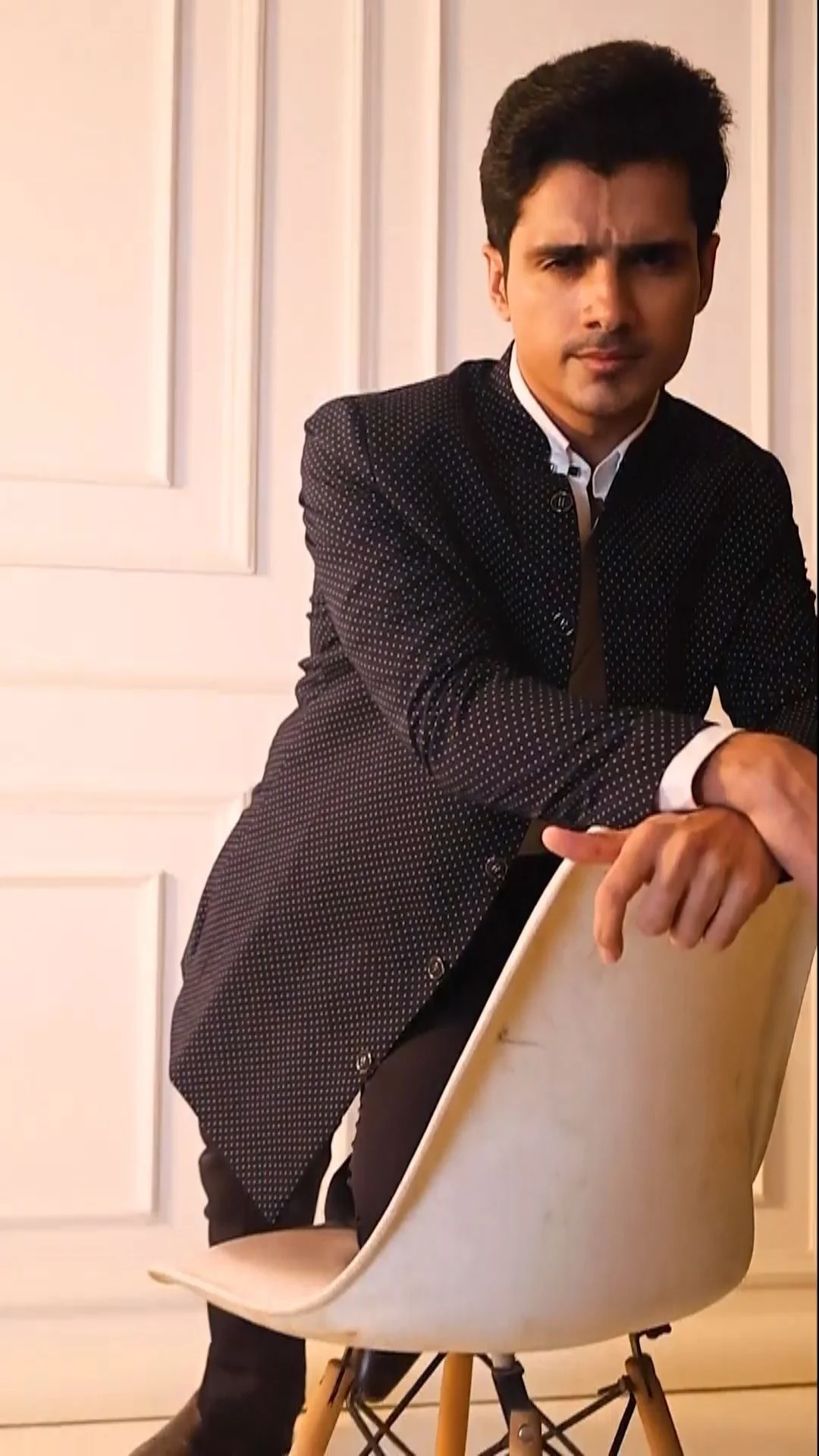 Indian male model in a dark textured blazer and white shirt seated backwards on a modern white chair in a bright panelled studio, menswear shoot where the commercial image consent covers stills and video