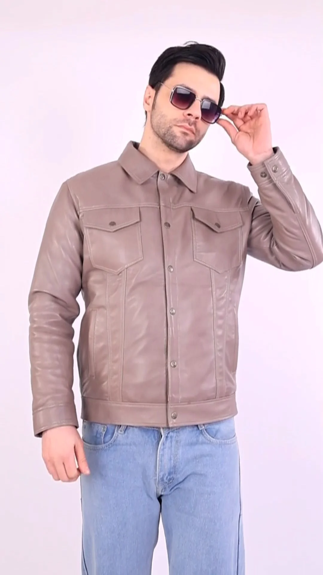 An Indian male model in a taupe leather jacket and blue jeans adjusts aviator sunglasses against a white studio wall, a styled fashion frame that hides the eyes and cannot replace unedited model photos