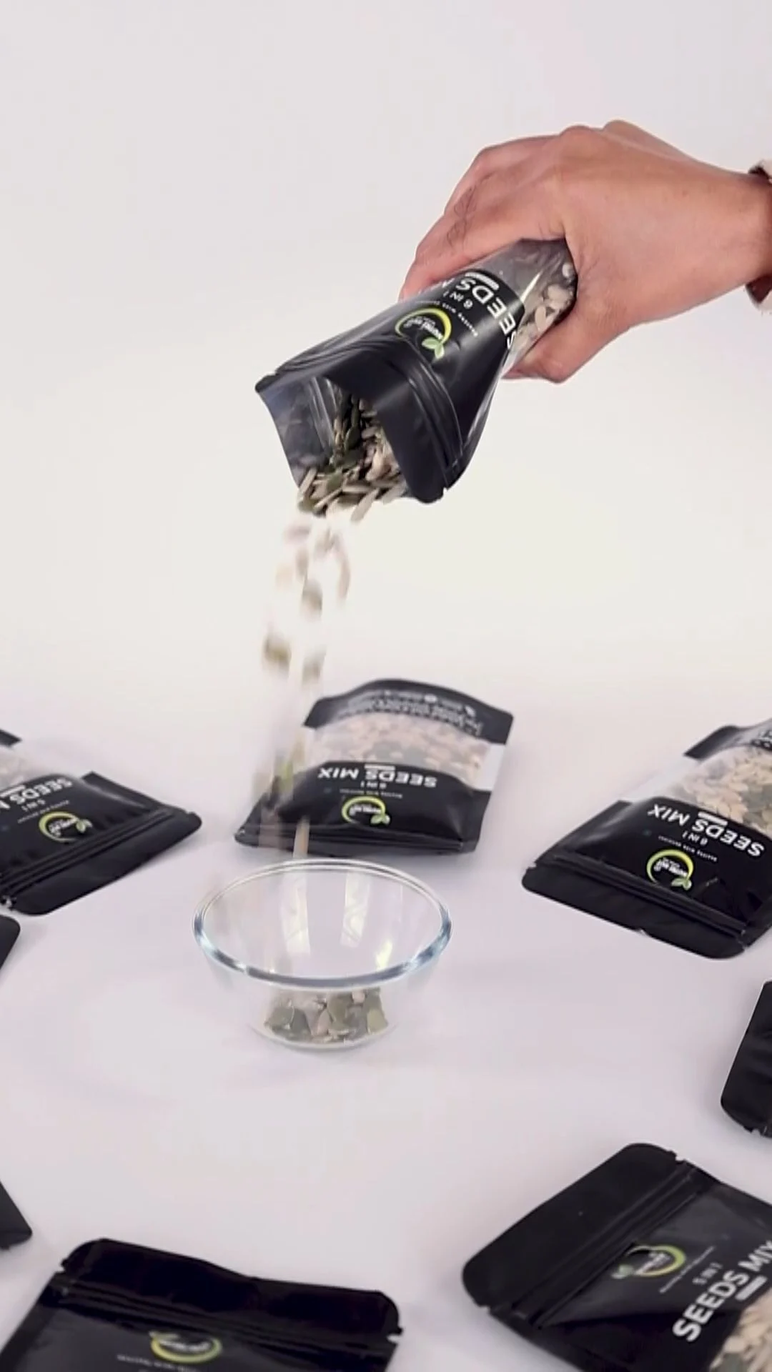 Hand pouring mixed dry fruits from a black pouch into a clear glass bowl on a white table surrounded by sealed pouches, a food product shot where claims and disclosure clauses matter as much as the usage term