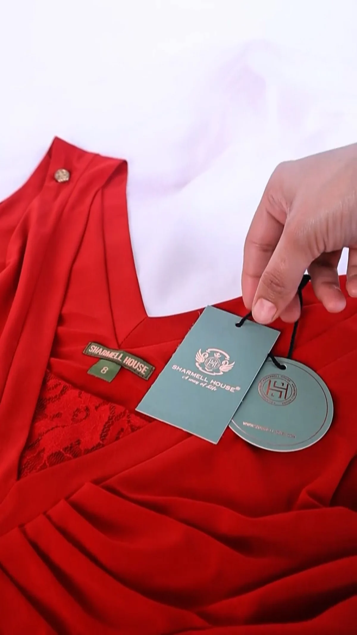 Close vertical detail shot of a hand lifting the swing tags on a red wrap dress laid on a white surface, the kind of product detail beat a D2C creator brief should list