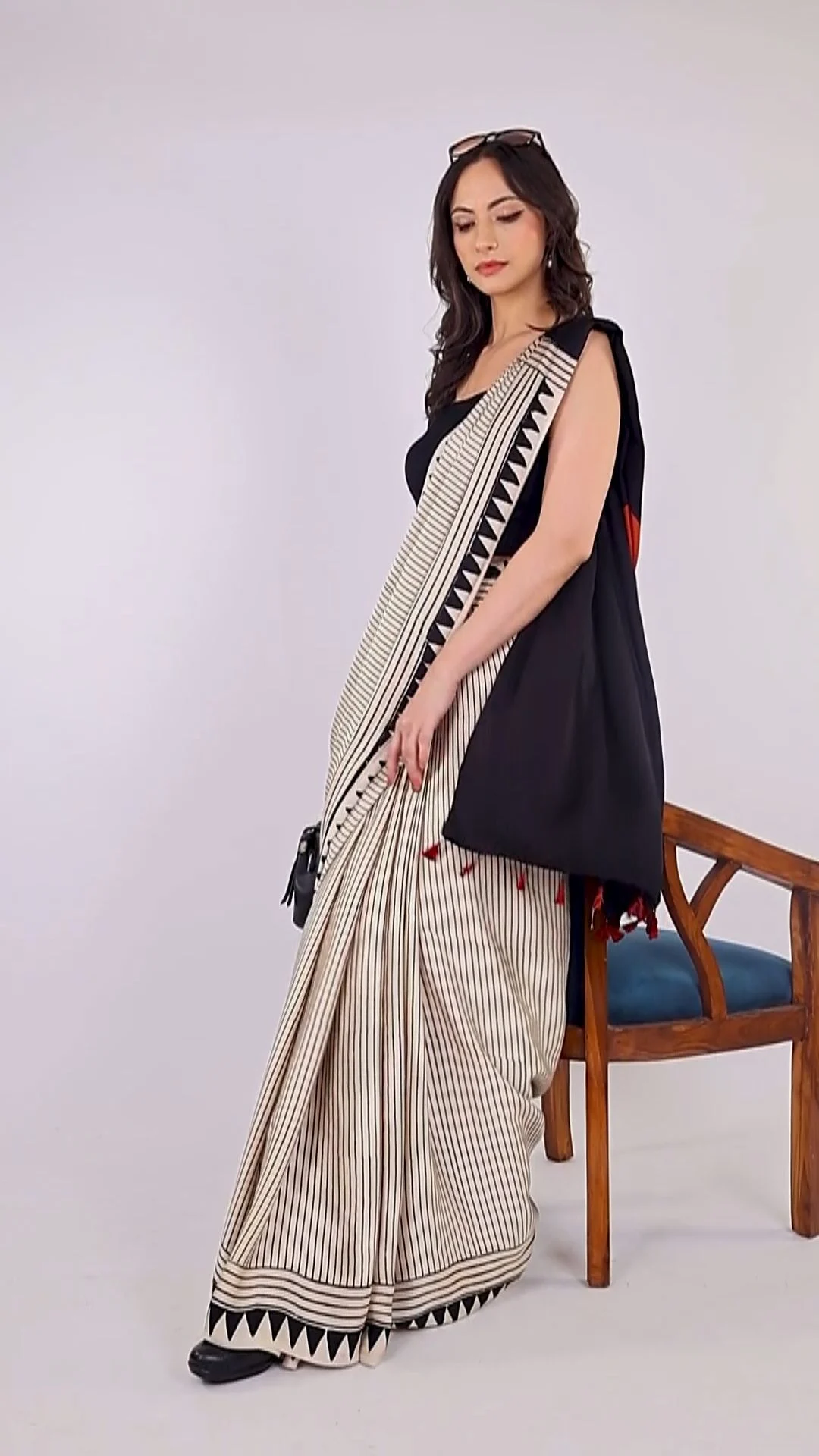 An Indian model in a striped cream handloom saree with a black border poses beside a wooden chair against a plain studio wall, an ethnic look for beginner model portfolio shots