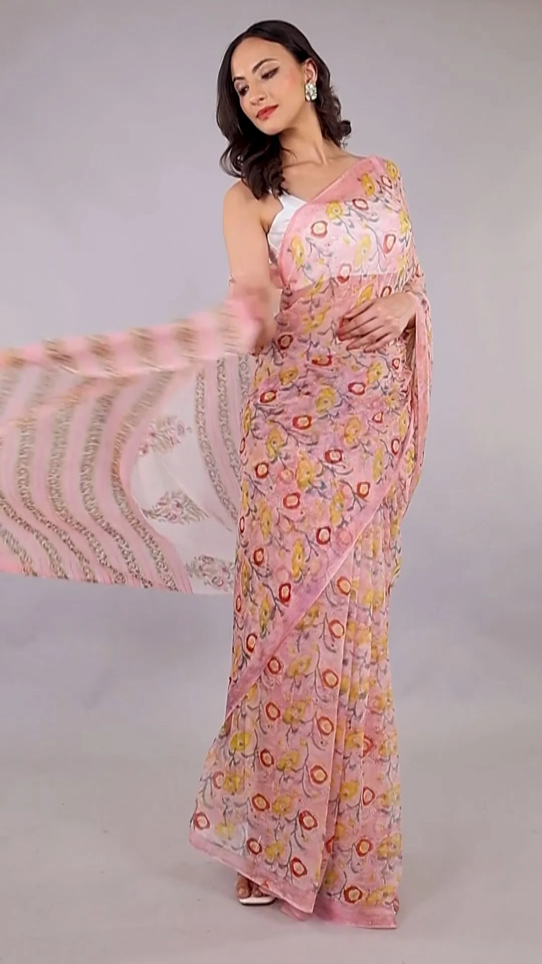 Indian woman model in a pink printed saree holding the pallu out to the side on a grey studio backdrop to show drape and border detail in a saree catalogue shoot