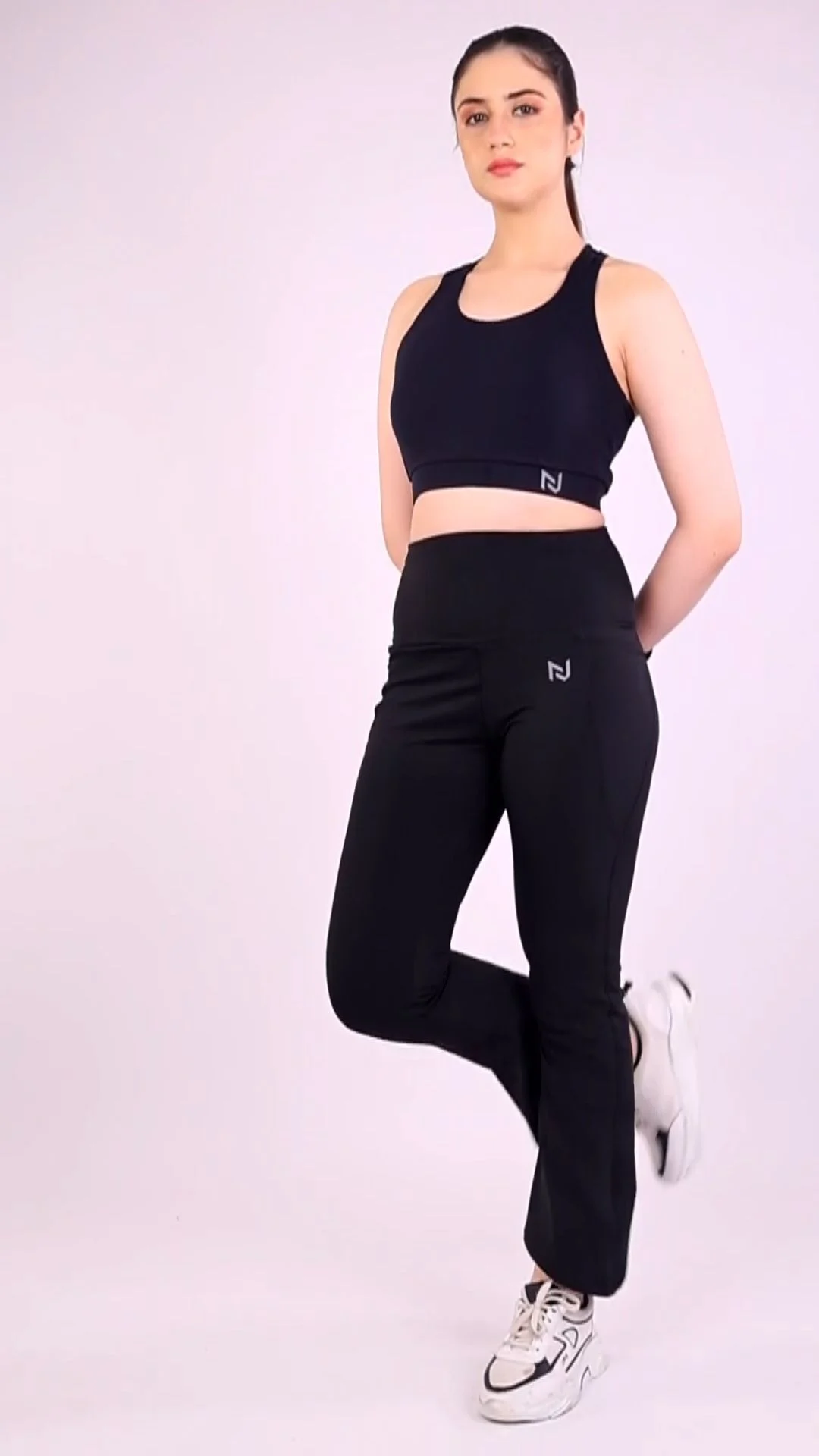 Full-length three-quarter view of an Indian woman in a fitted black sports bra and flared track pants against a pale lavender wall, showing the kind of fitted clothing that suits unedited model photos
