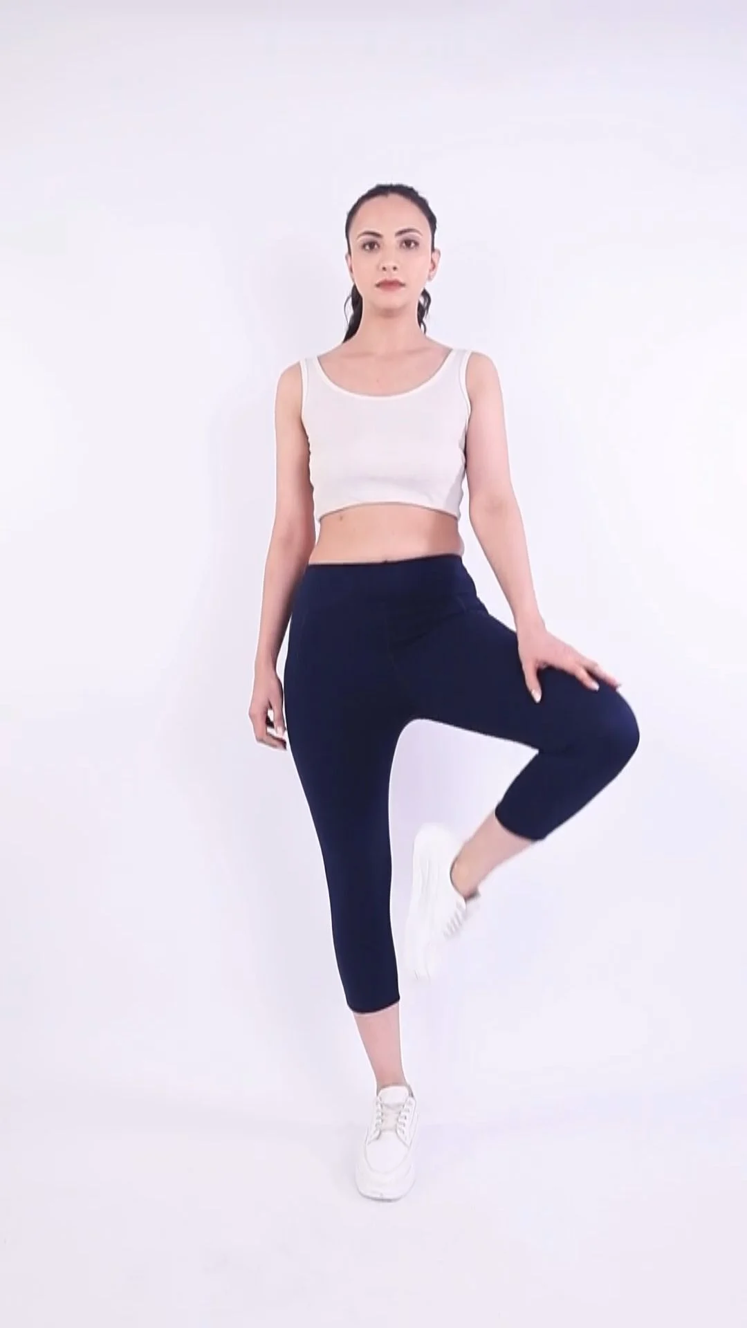 Indian female model in a white crop top, navy leggings and white sneakers balancing in a yoga pose against a seamless white studio backdrop, activewear catalogue image needing clear model usage rights India